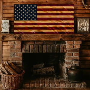 May include: A wooden American flag with red and tan stripes and a blue field of white stars hangs above a brick fireplace. A clock and a sign that says "hello honey" are also on the mantle. A basket of firewood and a kettle are in the foreground.