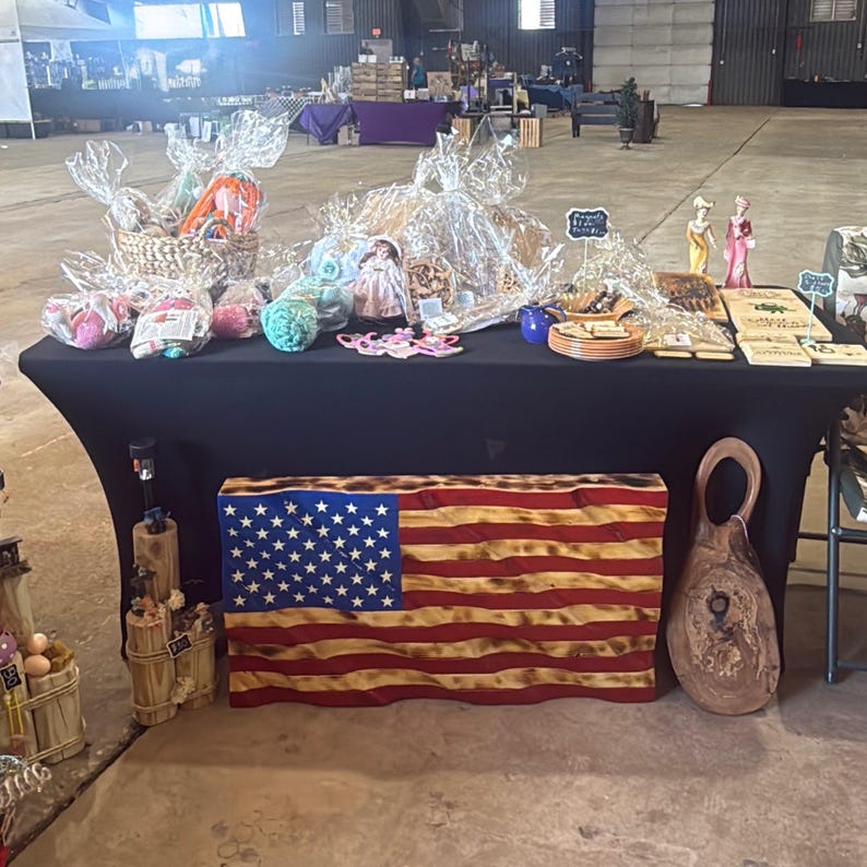 May include: A display table at a craft fair features various handmade items. A wooden American flag is prominently displayed, alongside wrapped gift baskets, decorative figurines, and wooden signs. The table is covered with a black cloth.