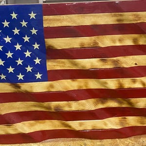 May include: Wooden American flag with a wavy design. The flag features a blue canton with white stars and alternating red and natural wood stripes. The wood has a distressed, rustic appearance.