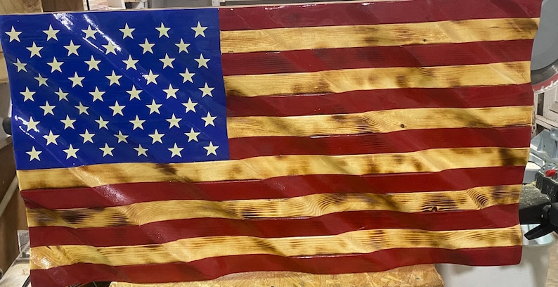 May include: Wooden American flag with a wavy design. The flag features a blue canton with white stars and alternating red and natural wood stripes. The wood has a distressed, rustic appearance.