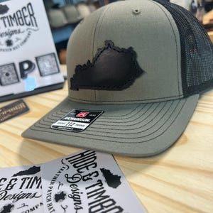 May include: A green and black trucker hat with a black leather patch of the state of Kentucky. The hat has the text "HIDE & TIMBER Designs" on the patch. The hat is on a wooden surface with two stickers that say "HIDE & TIMBER Designs" and "LEATHER PATCH HAT COMPANY".