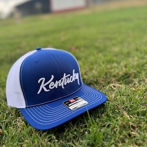 May include: A blue and white trucker hat with a white embroidered "Kentucky" logo. The hat has a mesh back and a white adjustable strap.