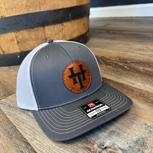 May include: Gray and white trucker hat with a brown leather patch featuring the letters "HH" in a circle. The hat has an adjustable strap with the text "Richardson 112" on it.