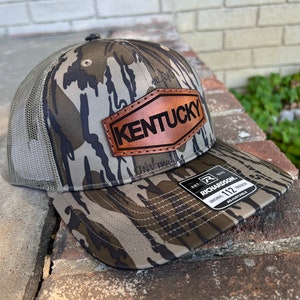 May include: A camouflage trucker hat with a brown leather patch that says "Kentucky" on it. The hat is made by Richardson 112.
