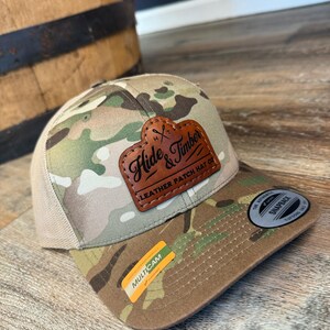 May include: A camouflage trucker hat with a brown leather patch that says "Hide & Timber Leather Patch Hat Co.". The hat has a mesh back and a brown brim.