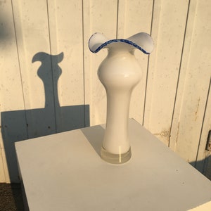 Op de afbeelding: Witte keramische vaas met een gerimpelde bovenkant en een blauwe rand. De vaas staat op een wit voetstuk.