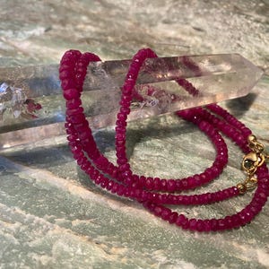 May include: A necklace composed of small, faceted, deep pink ruby beads strung together. The necklace features a gold-toned metal lobster claw clasp and a short extender chain, resting against a clear, elongated, hexagonal quartz crystal point.