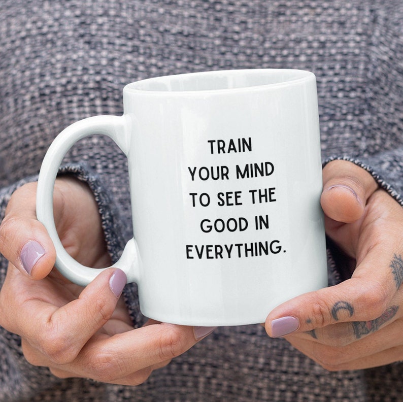 Op de afbeelding: Witte keramische mok met een zwart inspirerend citaat dat luidt: "Train je geest om het goede in alles te zien."