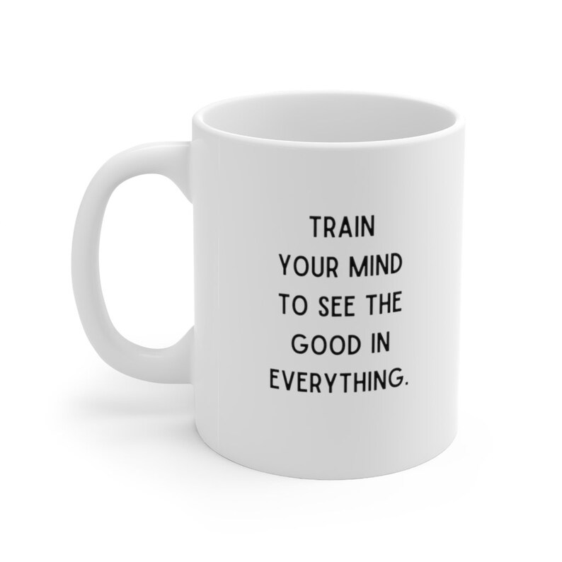 Op de afbeelding: Witte keramische koffiemok met zwarte tekst die luidt: "Train je geest om het goede in alles te zien."