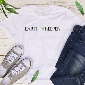 May include: White t-shirt with the text "EARTH KEEPER" and a small green leaf graphic. The shirt is laid out on a white wooden surface with a pair of gray sneakers and blue jeans.