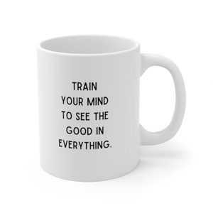 Op de afbeelding: Een witte keramische koffiemok met zwarte tekst die "Train je geest om het goede in alles te zien." zegt.