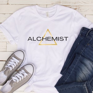 May include: White t-shirt with a gold triangle and the word "ALCHEMIST" in black text.