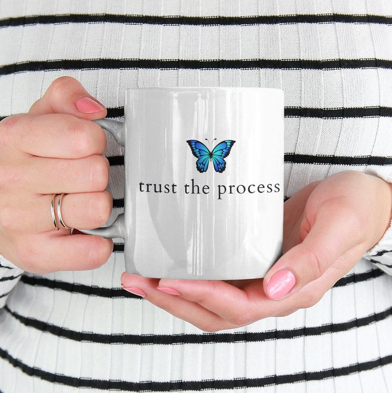 May include: White ceramic mug featuring a blue butterfly graphic and the text "trust the process" printed in black.