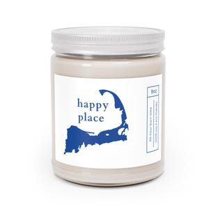 May include: A beige candle in a glass jar with a white lid. The label features the words "happy place" above a blue outline of a peninsula. The candle is 9oz and has a 50-hour burn time.