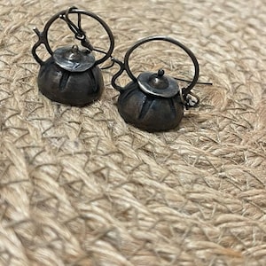 May include: A pair of silver teapot earrings with a detailed design. The earrings are hanging from a woven background.
