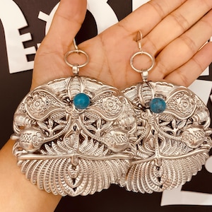May include: A pair of large, silver earrings with intricate floral designs and turquoise accents. The earrings are dangling and have a hook fastening.
