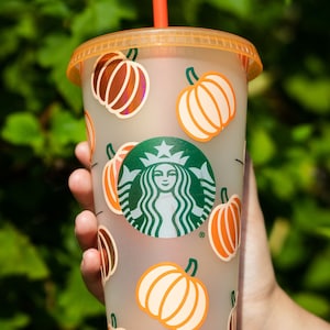 May include: A clear plastic reusable cup with a brown lid and a green Starbucks logo. The cup is decorated with orange and white pumpkin designs.