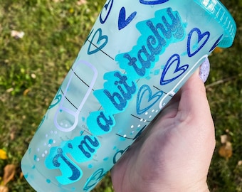 Pode incluir: Um copo reutilizável azul claro com uma tampa branca e um canudo azul. O copo tem um design de corações e o texto "I'm a bit tacky" em uma fonte de brilho azul.