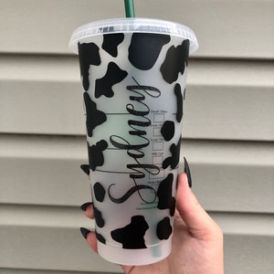 May include: A clear reusable cup with a black and white cow print design. The cup has a black lid and a green straw. The word "Sydney" is written on the side of the cup.