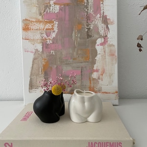 Könnte beinhalten: Zwei Keramikvasen in Form von menschlichen Hüften, eine schwarz und eine weiß. Die schwarze Vase hat getrocknete Blumen darin. Die Vasen stehen auf einem Buch mit dem Wort "JACQUEMUS" auf dem Einband. Das Buch liegt auf einer weißen Oberfläche.