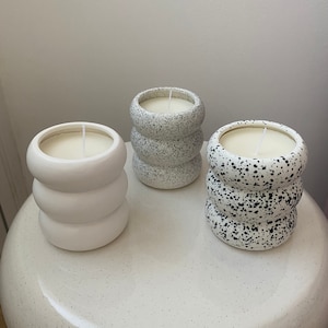 Könnte beinhalten: Drei weiße Kerzen aus Keramik mit gesprenkeltem Finish. Die Kerzen haben die Form eines Zylinders mit drei erhabenen Ringen. Die Kerzen sind in einer Reihe auf einer weißen Oberfläche angeordnet.