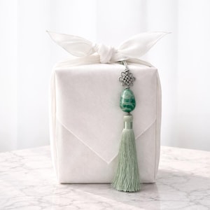 May include: A white fabric-wrapped gift box tied with a bow, adorned with a silver-toned charm and a green stone pendant. A matching green tassel hangs below the pendant, adding a touch of elegance.