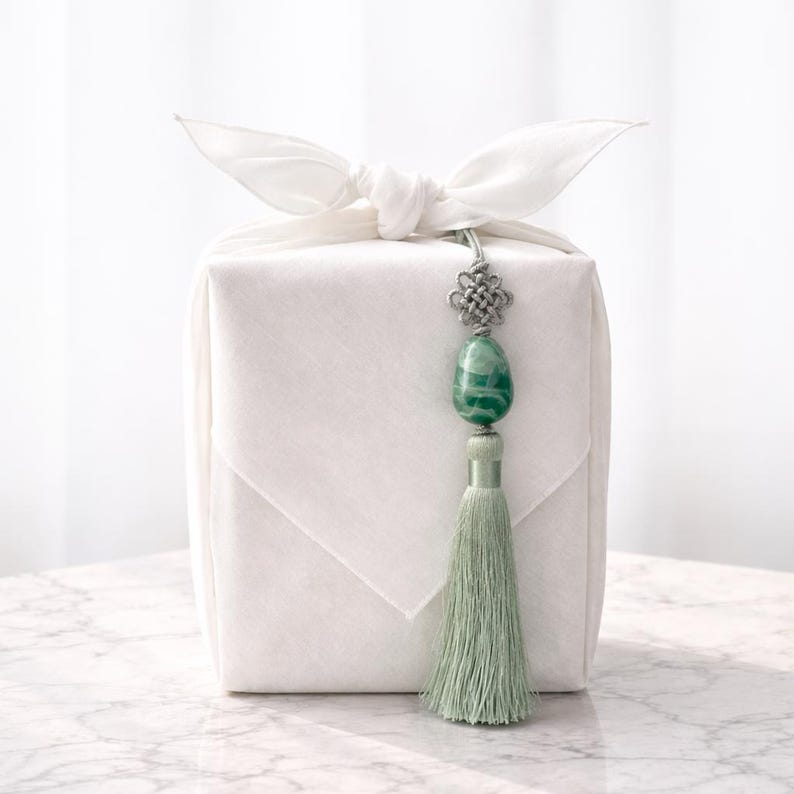 May include: A white fabric-wrapped gift box tied with a bow, adorned with a silver-toned charm and a green stone pendant. A matching green tassel hangs below the pendant, adding a touch of elegance.