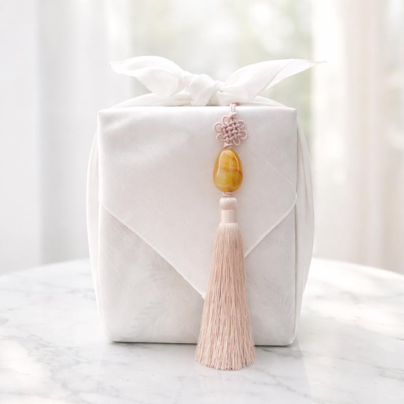 May include: A white gift box wrapped in fabric, tied with a bow, and adorned with a decorative tassel. The tassel is a light pink color, and the box has a decorative stone and knot detail.