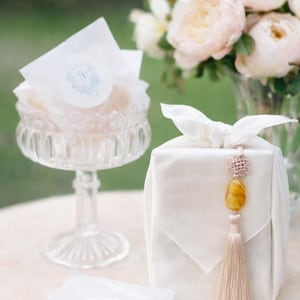 May include: A clear glass pedestal bowl filled with wrapped treats, a white fabric-wrapped gift with a decorative tassel, and a vase of pink flowers. The treats are wrapped in white paper with a blue and white emblem.