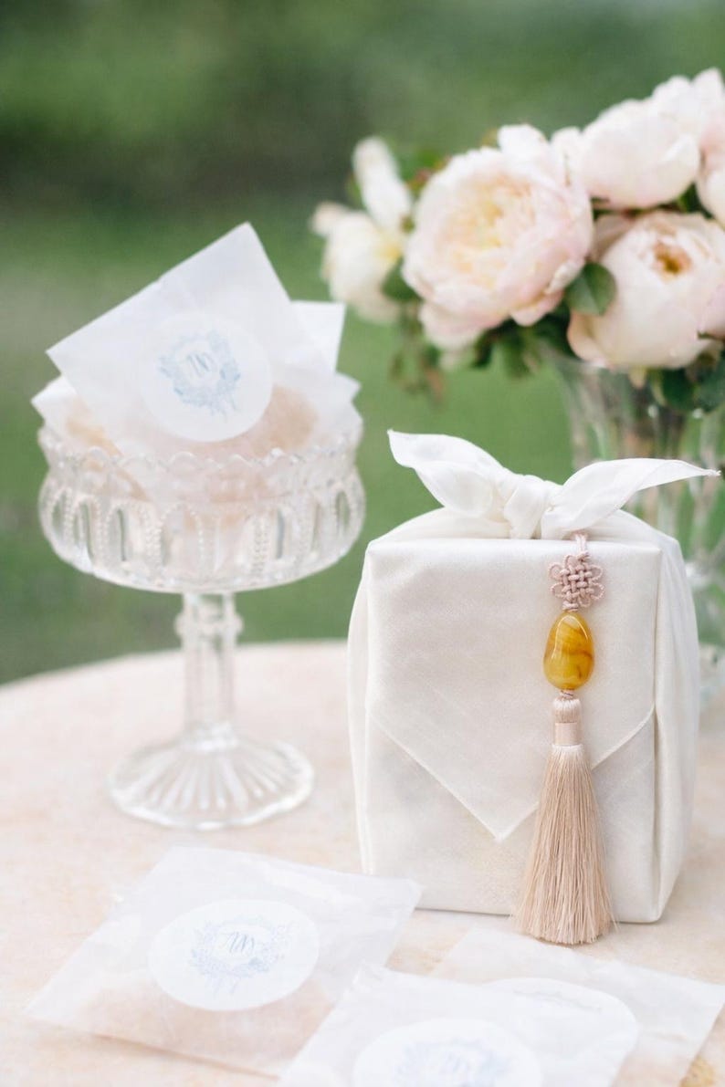 May include: A clear glass pedestal bowl filled with wrapped treats, a white fabric-wrapped gift with a decorative tassel, and a vase of pink flowers. The treats are wrapped in white paper with a blue and white emblem.