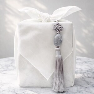 May include: A white, square gift box wrapped in fabric, tied with a bow, and adorned with a gray tassel and decorative knot. The tassel hangs from a gray stone pendant. The overall aesthetic is minimalist and elegant.