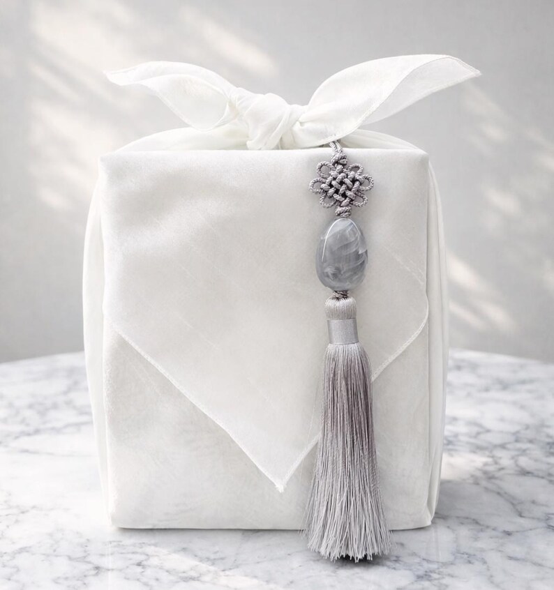 May include: A white, square gift box wrapped in fabric, tied with a bow, and adorned with a gray tassel and decorative knot. The tassel hangs from a gray stone pendant. The overall aesthetic is minimalist and elegant.