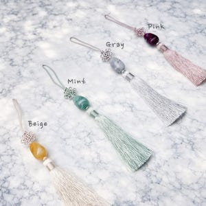 May include: Four decorative tassels in beige, mint, gray, and pink. Each tassel features a gemstone bead, a silver-colored knot, and a matching silky tassel. The colors are labeled on the image.