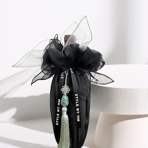 May include: A black wine bottle bag with a decorative bow made of sheer black and silver fabric. The bag is adorned with a black ribbon that reads "STYLE BY HM" and a jade green tassel. The bottle is on a white surface.