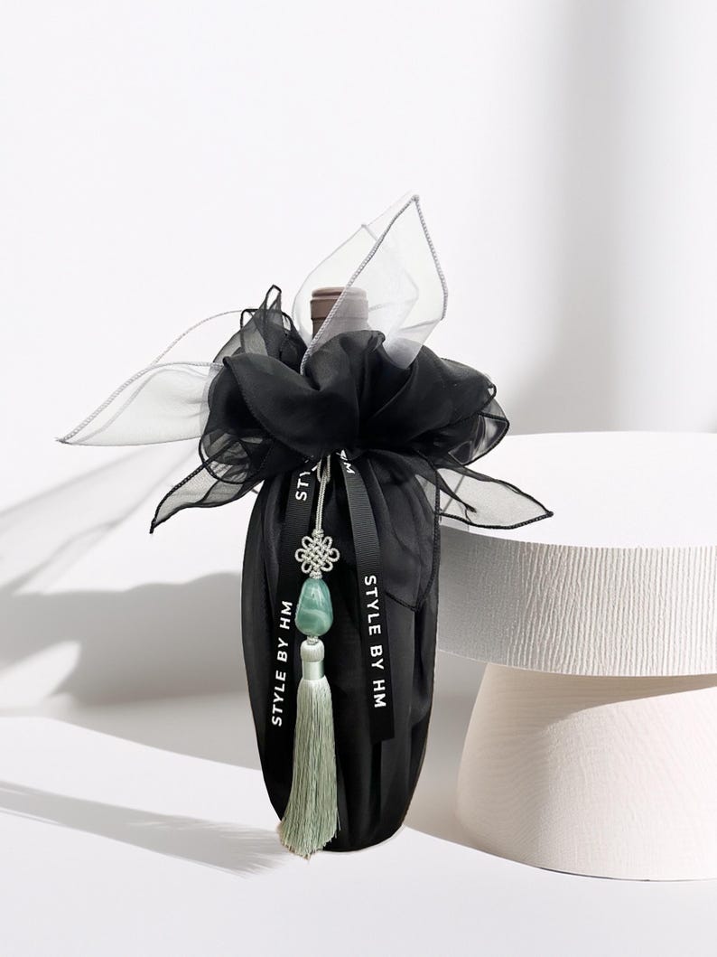 May include: A black wine bottle bag with a decorative bow made of sheer black and silver fabric. The bag is adorned with a black ribbon that reads "STYLE BY HM" and a jade green tassel. The bottle is on a white surface.