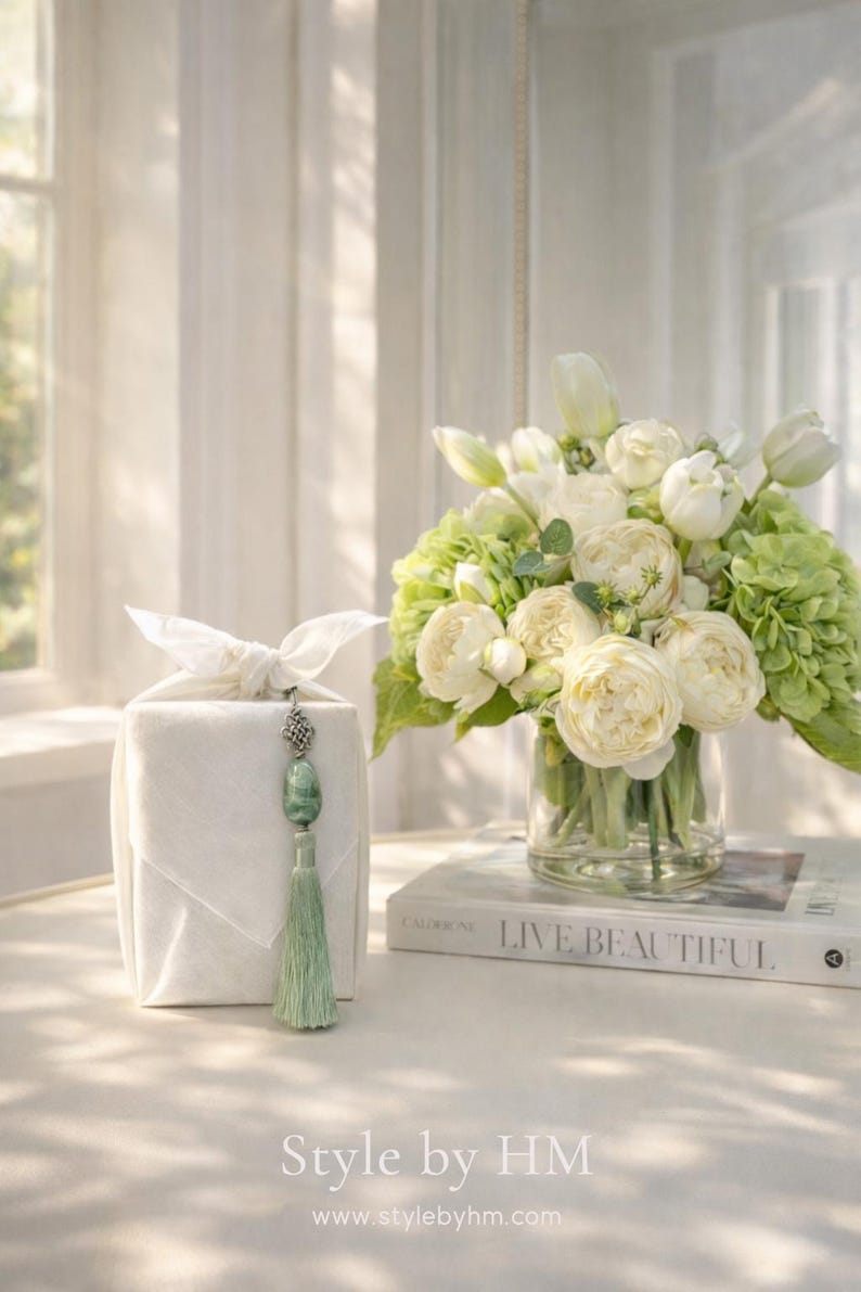 May include: A white gift box with a decorative tassel and a bouquet of white roses, tulips, and green hydrangeas in a glass vase. The book "LIVE BEAUTIFUL" is visible. The text "Style by HM" and website address are at the bottom.