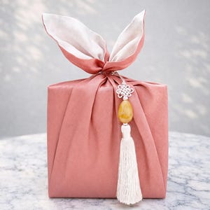 May include: A wrapped gift in a dusty rose fabric, tied with a bow that resembles bunny ears. A decorative charm with a yellow stone and white tassel hangs from the knot. The gift is presented on a marble surface.