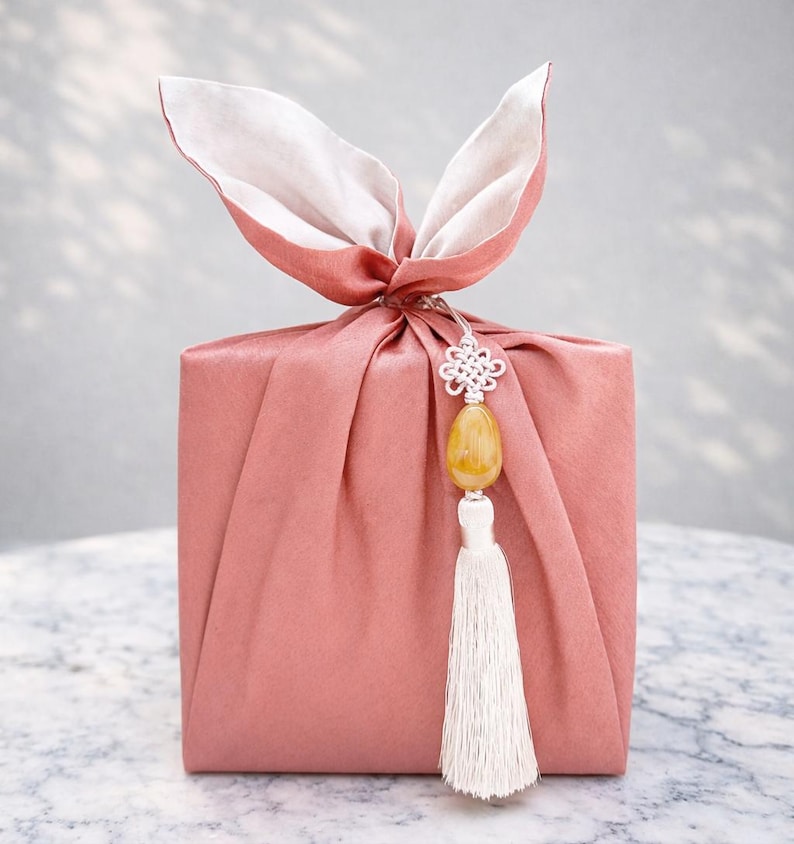 May include: A wrapped gift in a dusty rose fabric, tied with a bow that resembles bunny ears. A decorative charm with a yellow stone and white tassel hangs from the knot. The gift is presented on a marble surface.