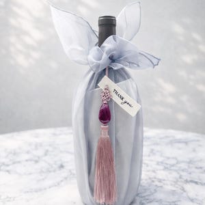 May include: A wine bottle wrapped in light blue fabric and tied with a matching bow. A tag that says "Thank you" hangs from a decorative tassel with a purple gem and pink tassel.