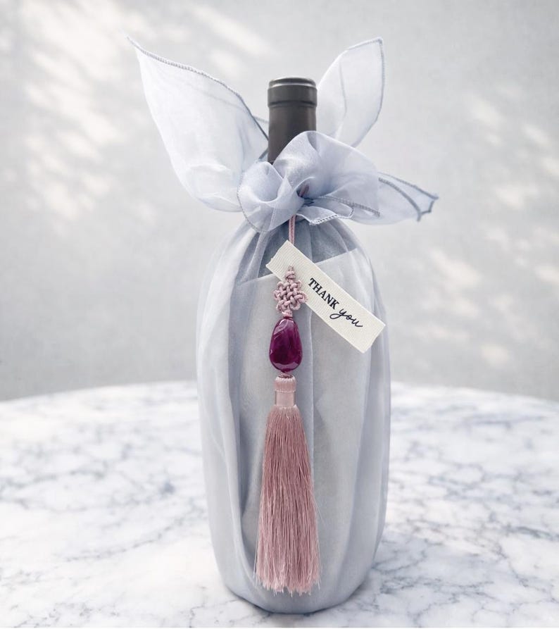 May include: A wine bottle wrapped in light blue fabric and tied with a matching bow. A tag that says "Thank you" hangs from a decorative tassel with a purple gem and pink tassel.
