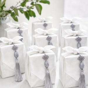 May include: White gift boxes wrapped in fabric, tied with white bows, and adorned with gray tassels and decorative ornaments. The gifts are arranged on a white surface, with greenery in the background.