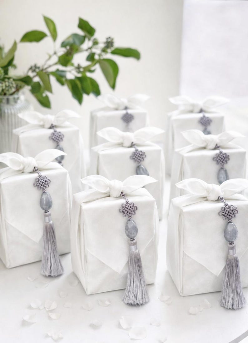 May include: White gift boxes wrapped in fabric, tied with white bows, and adorned with gray tassels and decorative ornaments. The gifts are arranged on a white surface, with greenery in the background.