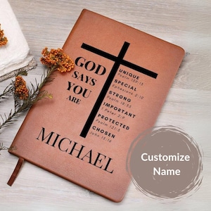 Puede incluir: Un diario de cuero marr&oacute;n con una cruz y el texto "GOD SAYS YOU ARE" y una lista de vers&iacute;culos b&iacute;blicos. El nombre "MICHAEL" est&aacute; impreso en la parte inferior del diario. El texto "Customize Name" est&aacute; impreso en un c&iacute;rculo en el lado derecho del diario.