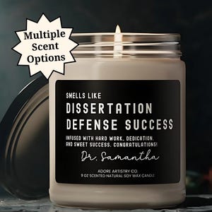 May include: A brown soy wax candle in a clear glass jar with a black label that reads "SMELLS LIKE DISSERTATION DEFENSE SUCCESS INFUSED WITH HARD WORK, DEDICATION, AND SWEET SUCCESS. CONGRATULATIONS! Dr. Samantha ADORE ARTISTRY CO. 9 OZ SCENTED NATURAL SOY WAX CANDLE".