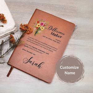 May include: A brown leather journal with a floral design and the text "Difference Maker" defined as "a person who accomplishes extraordinary things by creating positive change in their environment and in the lives of others." The journal also has the text "Also known as: Sarah" and "Customize Name".