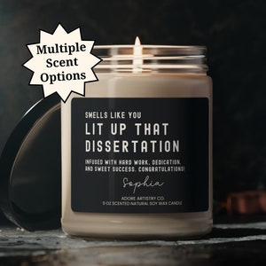 May include: A black soy wax candle with a white label that reads "Smells like you Lit up that Dissertation. Infused with hard work, dedication, and sweet success. Congratulations! Sophia. Adore Artistry Co. 9 oz Scented Natural Soy Wax Candle".