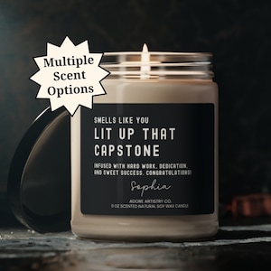 May include: A black and white candle jar with the text "Smells Like You Lit Up That Capstone" and "Infused with hard work, dedication, and sweet success. Congratulations!"  The candle jar is labeled "Sophia" and "Adore Artistry Co. 9 oz Scented Natural Soy Wax Candle".