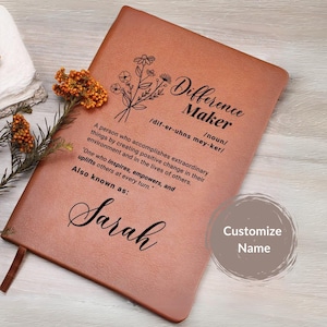May include: A brown leather journal with the words "Difference Maker" defined as "a person who accomplishes extraordinary things by creating positive change in their environment and in the lives of others." The journal also has a floral design and the name "Sarah" written in cursive.