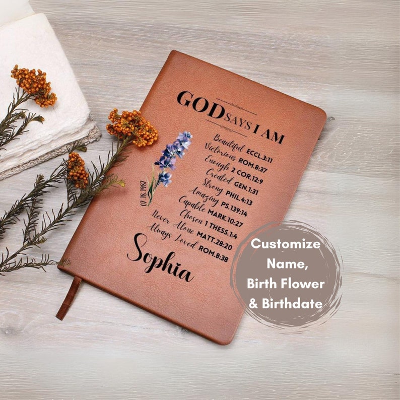 Puede incluir: Un diario de cuero marr&oacute;n con el texto "GOD SAYS I AM" y una lista de afirmaciones positivas. El diario est&aacute; decorado con un dise&ntilde;o floral y el nombre "Sophia" est&aacute; escrito en la parte inferior. El texto "Customize Name, Birth Flower & Birthdate" tambi&eacute;n est&aacute; incluido.