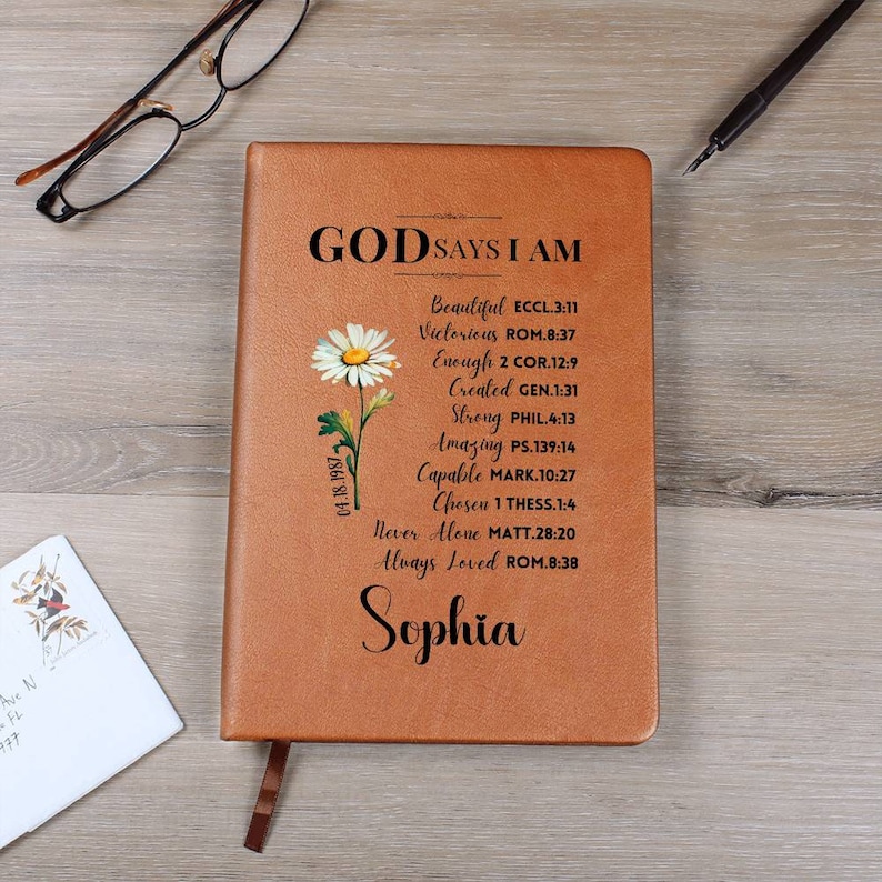 Puede incluir: Un diario de cuero marr&oacute;n con las palabras "God Says I Am" y un dise&ntilde;o de margarita. El diario tiene una lista de afirmaciones positivas con vers&iacute;culos b&iacute;blicos. El nombre "Sophia" est&aacute; escrito en la parte inferior.